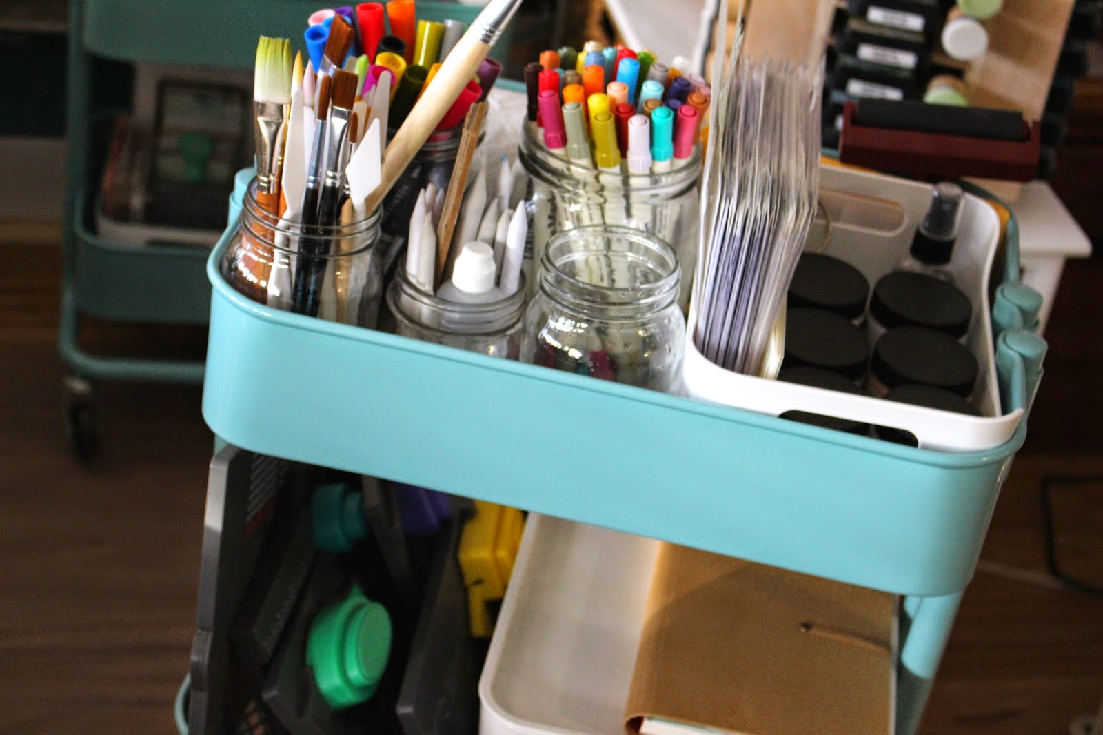 A teal craft cart holding jars of paintbrushes, colored markers, and other art supplies, with additional storage visible